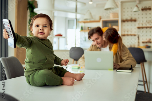 Portrait of a baby sitting on the table and playing with a calculator while his father is trying to work, in the behind. photo