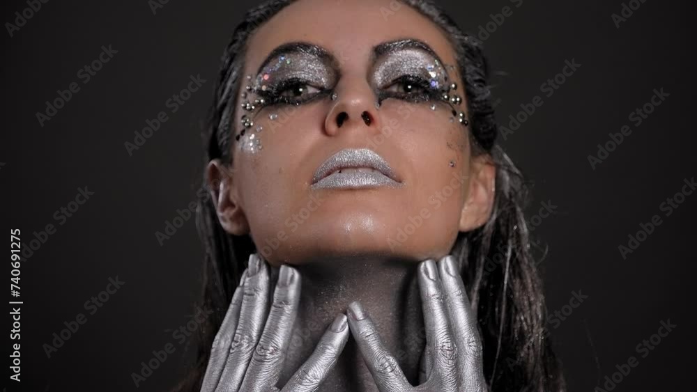 Close-up of a gorgeous female model with silver skin and hands, with bright makeup and stones, she poses in the studio on a black background. Fashion portrait of a young woman. Close-up of a gorgeous female model with silver skin and hands, with bright makeup and stones, she poses in the studio on a black background. Fashion portrait of a young woman.