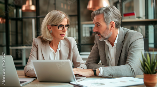 Senior Business Colleagues Working Together on Laptop. A focused senior man and woman in professional attire working closely on a laptop in a modern office setting.