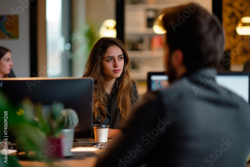 Focused Young Female Professional in Office Meeting. A young woman attentively listens during a business meeting, with a blurred male colleague in the foreground, in a modern office setting.