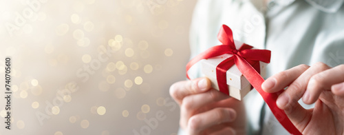Close-up of a woman holding a gift and tying a beautiful red ribbon bow, banner space for text