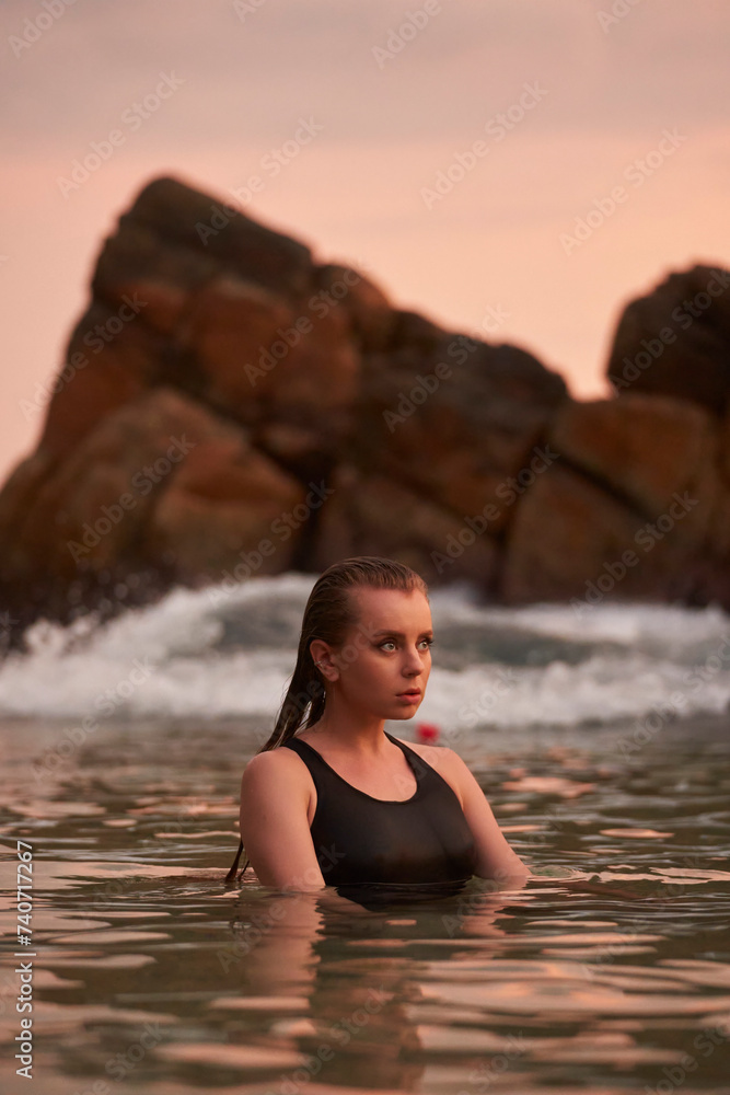 Fototapeta premium Woman relaxes in sea at sunset. Tranquil marine swim, wellness leisure activity. Fit female in swimsuit enjoys natural water, serenity at dusk. Harmony with ocean, healthy beach lifestyle. Solo travel