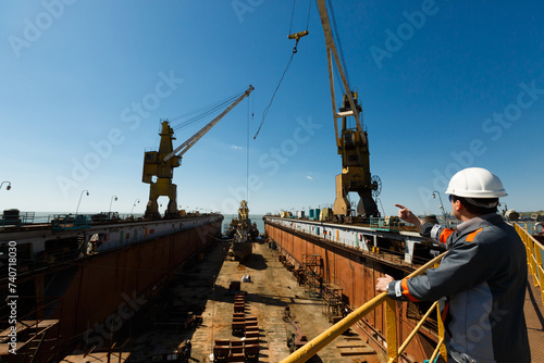 Engineer oversees vessel maintenance in dry dock. Maritime worker points, directs crane at shipyard under sunny sky. Industrial worker inspects, guides ship overhaul. photo