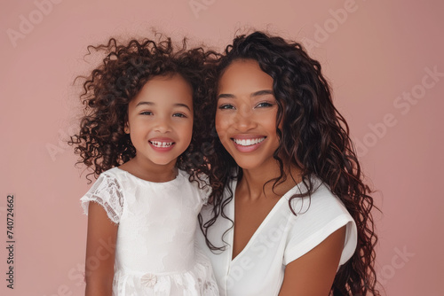 Pareja de hermanas afroamericanas de diferentes edades  sonrientes de pelo largo rizado, con vestido blanco, sobre fondo rosa claro pastel photo