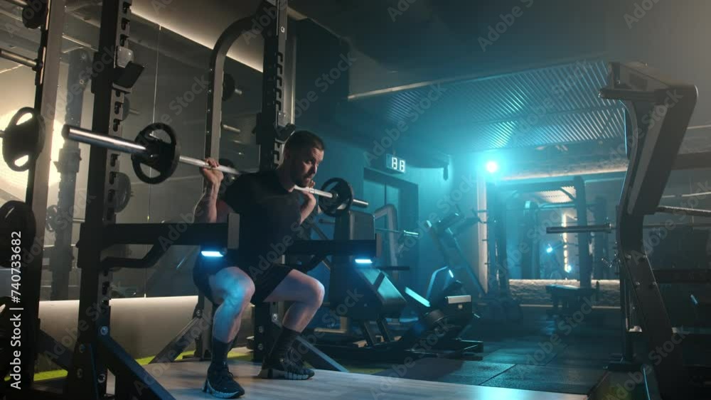 A focused man is captured mid-repetition doing barbell squats in a contemporary gym setting, illuminated by the stark blue lights that cast a cool hue over the well-equipped space.