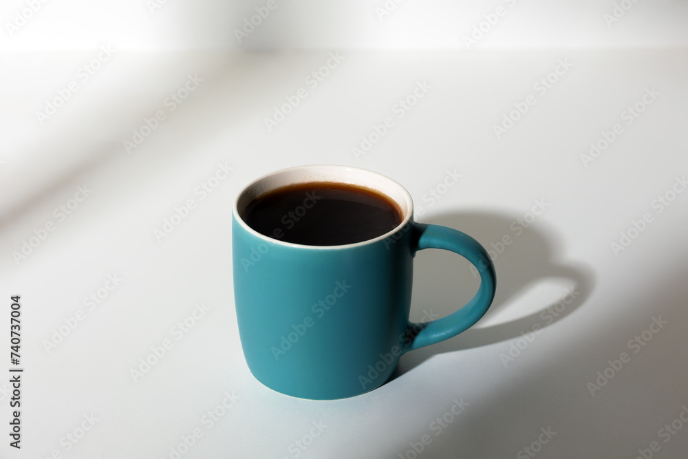 A cup of coffee stands on a white background with a shadow from the window in the morning light