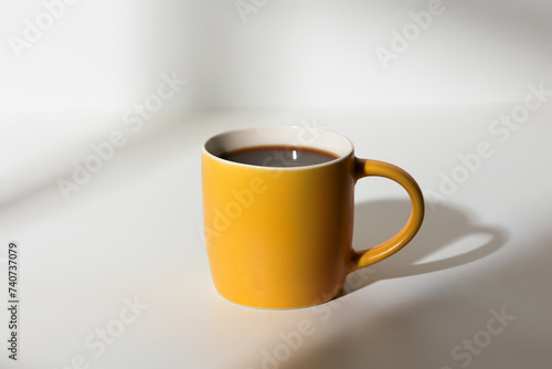 A cup of coffee stands on a white background with a shadow from the window in the morning light