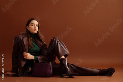 Fashionable confident woman wearing faux leather suit blazer, pants, pointed toe ankle boots, with trendy purple bag, posing on brown background. Full-length studio fashion portrait. Copy, empty space