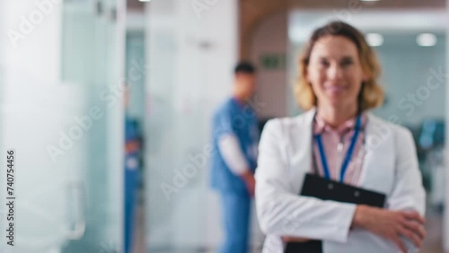 Female doctor wearing white lab coat and holding clipboard walks into focus in hospital corridor with colleagues in background - shot in slow motion photo
