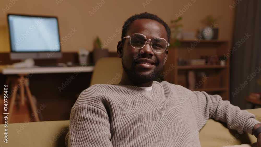 Zoom-in portrait of young Black man in eyeglasses sitting on comfortable soft couch in cozy living room and smiling at camera Zoom-in portrait of young Black man in eyeglasses sitting on comfortable soft couch in cozy living room and smiling at camera