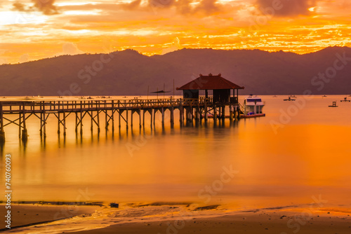 A wooden pier stretches out into a calm bay at sunset, with mountains in the background. © Neilstha Firman