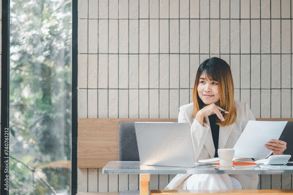 A confident businesswoman assesses documents with a pen in hand, laptop open, in the bright ambiance of a cafe setting. A confident businesswoman assesses documents with a pen in hand, laptop open, in the bright ambiance of a cafe setting.