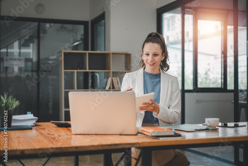 Obraz na plátne Business analytics concept, A smiling young professional woman using a laptop and holding a notebook in a bright, modern office setting
