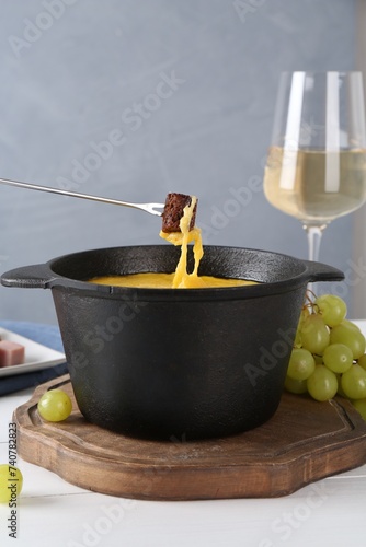 Dipping piece of bread into fondue pot with tasty melted cheese at white wooden table against gray background  closeup