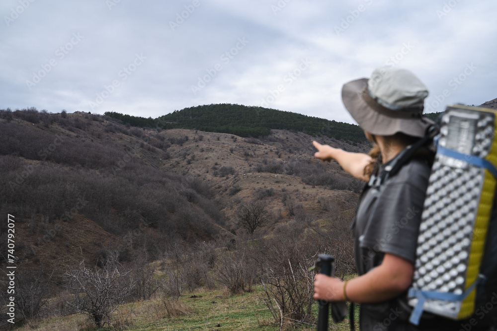 Back view of young female hiker with tourist gears and trekking poles points with finger at the mountains background, camping journey tourism adventure concept, focus on the mountains, copy space Back view of young female hiker with tourist gears and trekking poles points with finger at the mountains background, camping journey tourism adventure concept, focus on the mountains, copy space