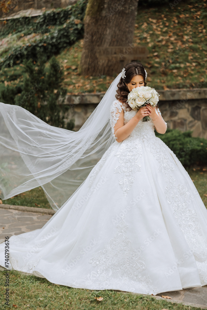 A brunette bride in a voluminous white dress holds a bouquet, poses. The veil is thrown into the air. Beautiful hair and makeup. Wedding photo session in nature.