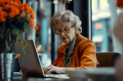 An elegant elderly lady in a yellow blazer attentively uses a computer in a cozy cafe setting with vibrant flowers. 