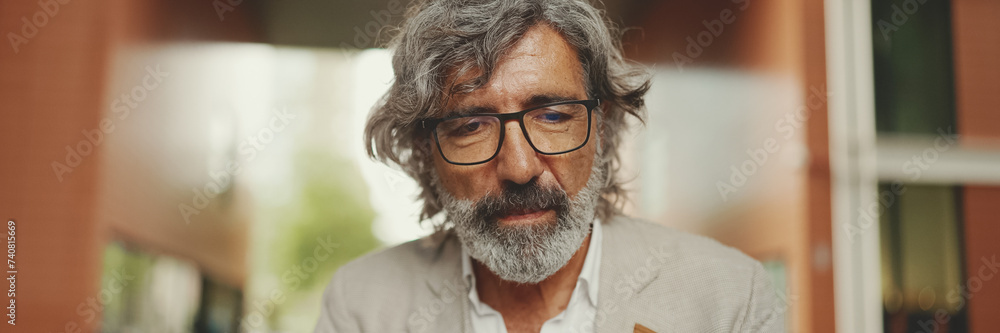 Clouse up, mature businessman with beard in eyeglasses wearing gray jacket sits on cafe, Panorama. Middle aged manager successful man user on laptop pc computer sit at cafe outdoors Clouse up, mature businessman with beard in eyeglasses wearing gray jacket sits on cafe, Panorama. Middle aged manager successful man user on laptop pc computer sit at cafe outdoors