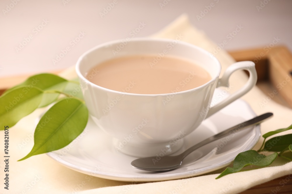 Cup of aromatic tea with milk, spoon, saucer and green leaves on table, closeup Cup of aromatic tea with milk, spoon, saucer and green leaves on table, closeup