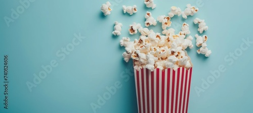 Popcorn spilling from red striped box on pastel blue background, with space for text, food concept.