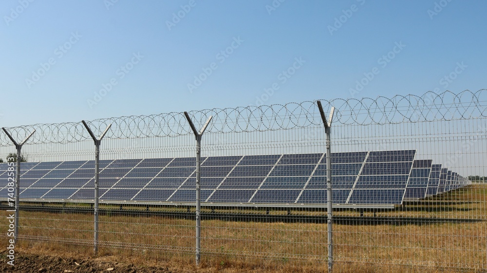 a complex of solar panels behind a barbed wire fence in a field under a clear blue sky, a country solar power station behind a guarded barrier, solar energy in industry a complex of solar panels behind a barbed wire fence in a field under a clear blue sky, a country solar power station behind a guarded barrier, solar energy in industry