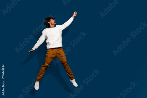 Full length photo of cheerful positive man wear white sweater jumping high catching arm empty space isolated dark blue color background
