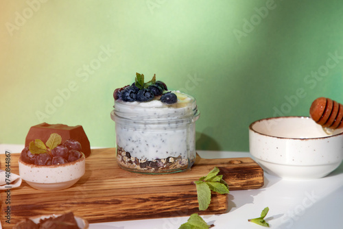 Yogurt in a glass jar with granola, chocolate, blueberries and honey on a wooden board on a green background with mint leaves. Front view