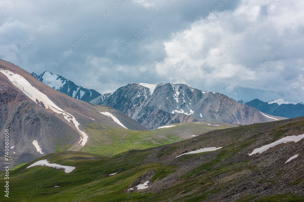Dramatic alpine view to big rocky ridge and stony green hills under rainy gray sky. Overcast landscape with large wide snow-capped mountain range. Beautiful hilly terrain and snowy mountains far away.