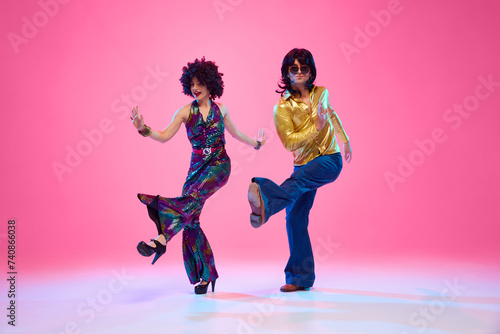 Fototapeta Naklejka Na Ścianę i Meble -  Retro disco era fashion. Couple with funky hairstyles and groovy clothes dancing energetic dance against gradient pink studio background. Concept of American culture, 1970s, 1980s fashion, music.