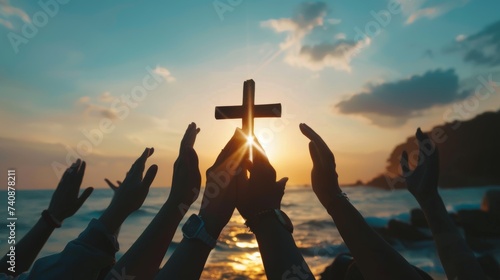In the background of a sunset, a group of hands is praying and holding a christian cross. Silhouette shows hands holding the cross as they worship God.