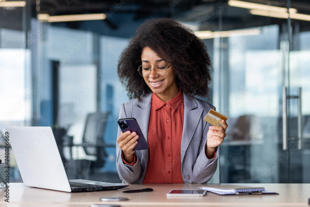 Satisfied african american female keeping debit card and cellular phone in hands by desk with computer and notebook. Pleased lady entering bank data in application for completing cashless payment Satisfied african american female keeping debit card and cellular phone in hands by desk with computer and notebook. Pleased lady entering bank data in application for completing cashless payment