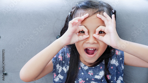 Young Asian girl over gray couch background doing ok gesture like binoculars, eyes looking through fingers.