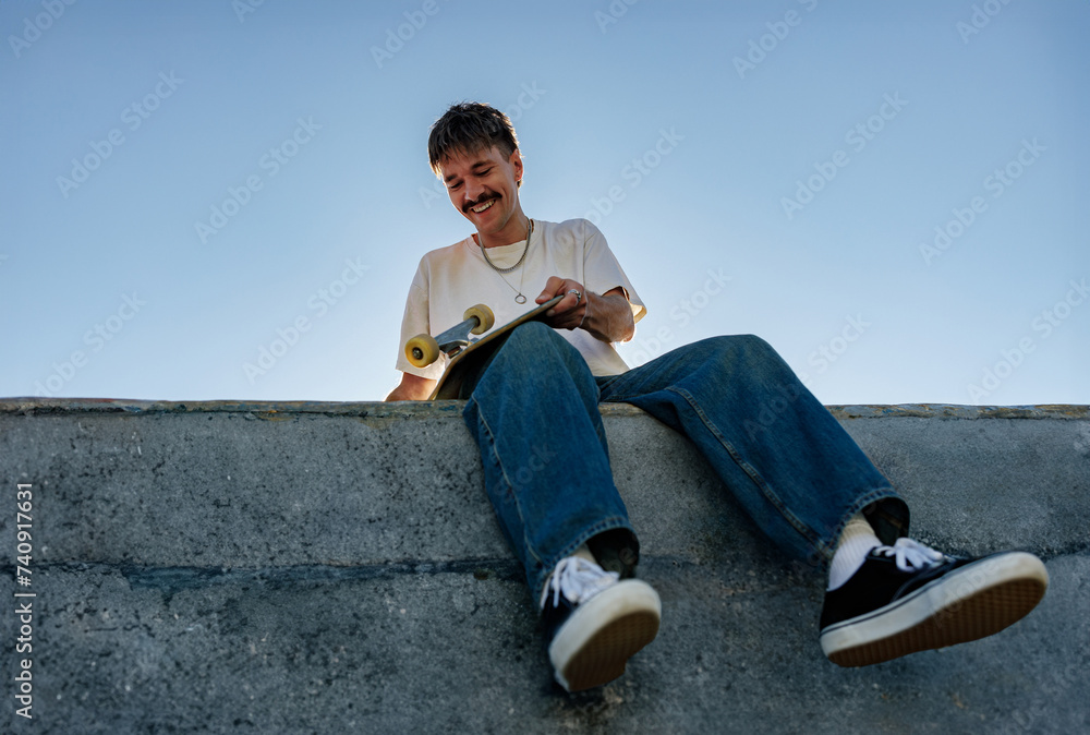 Low angle of young male skateboarder holding skateboard sitting on ramp in skate park 