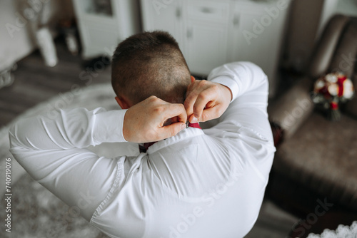 a young man in a white shirt stands by the window in the room and puts on a tie. The groom gets dressed in the morning and prepares for the wedding. © Vasil
