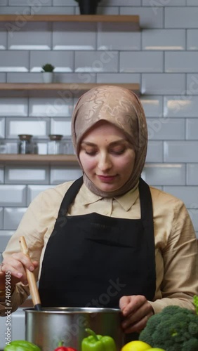 Vertical video. Beautiful young woman with headscarf cooks soup at home in modern kitchen and checks its taste	
