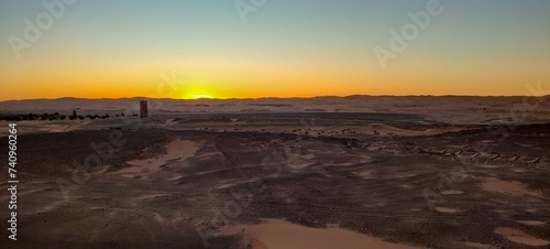 A sweeping panorama encapsulates the expansive sandy desert, featuring solitary vegetation on arid terrain, and a stunning array of rocks shaped by nature during the sunset in Timimoun, Algeria. photo