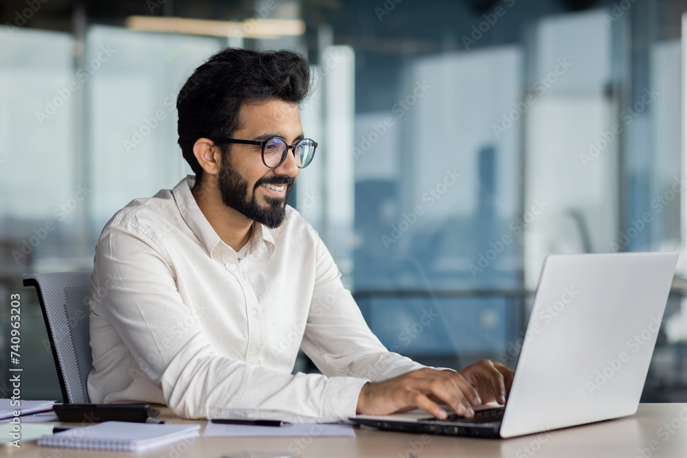 © Tetiana - A smiling young Indian man in a suit sits in the office at a desk and works on a laptop, typing on a keyboard, texting, working with data © Tetiana - A smiling young Indian man in a suit sits in the office at a desk and works on a laptop, typing on a keyboard, texting, working with data