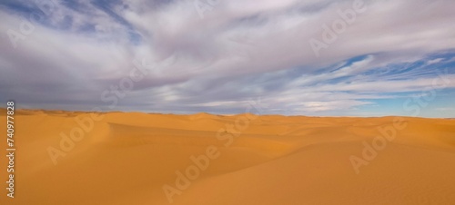 A sweeping panorama captures the vast expanse of golden sand dunes stretching as far as the eye can see  forming an arid desert under a partly cloudy blue sky in Timimoun  Algeria.