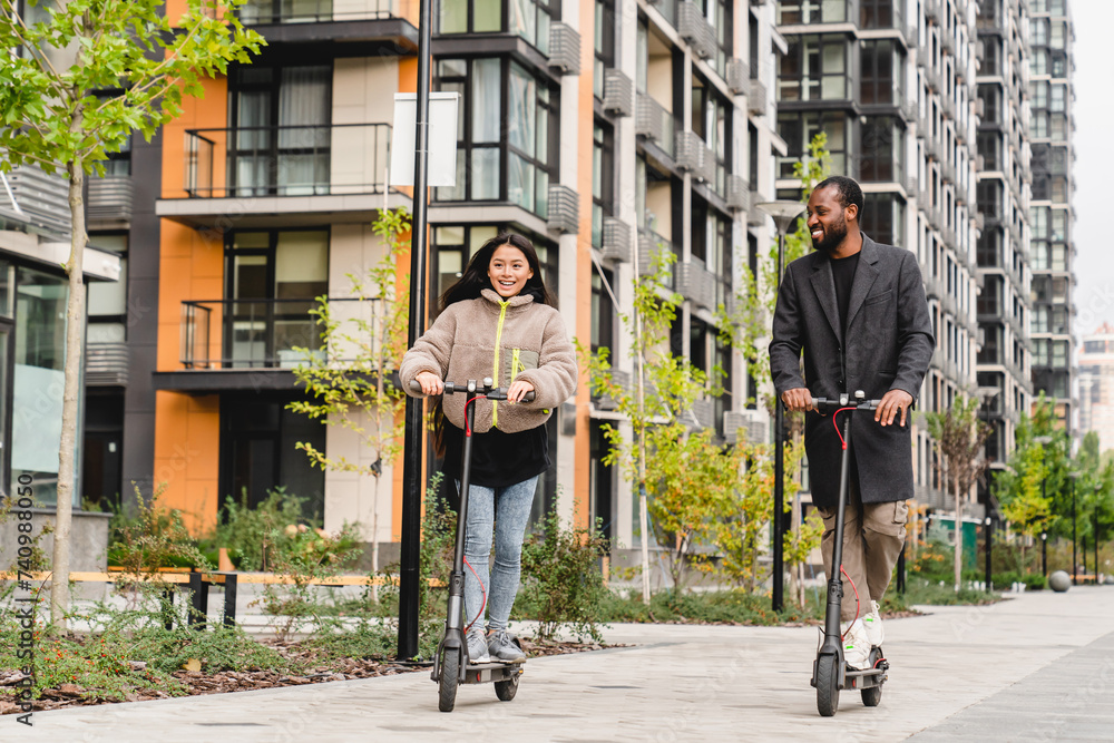 Young multiethnic couple riding electric scooters and talking on the street near modern buildings. Romantic date of young girlfriend and boyfriend on eco-transport outdoors