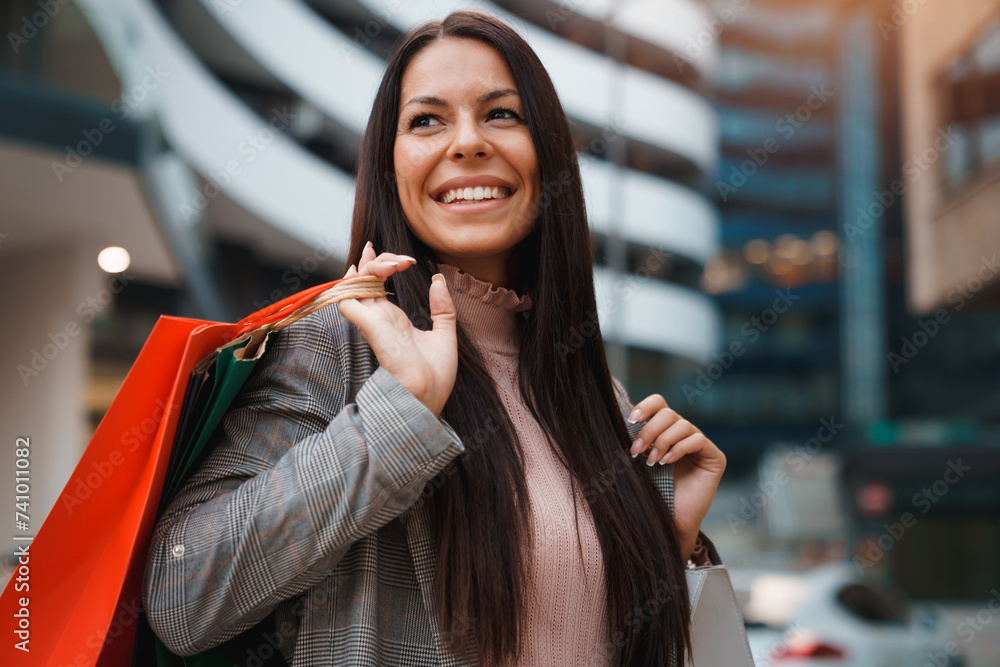 Portrait of pretty young woman with long straight hair standing in front of modern building and smiling while holding shopping bags. Portrait of pretty young woman with long straight hair standing in front of modern building and smiling while holding shopping bags.