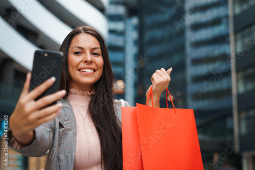 Happy smiling woman standing in front of modern city building and taking a selfie with shopping bag.