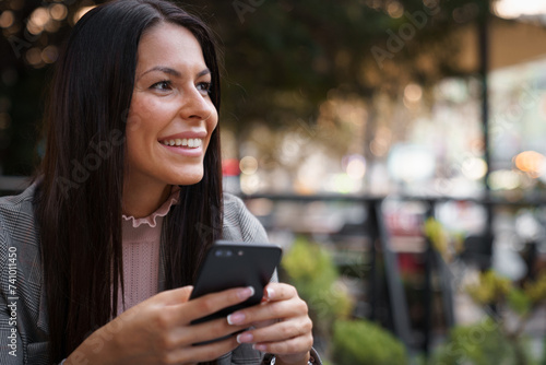 Portrait of beautiful brunette with long straight hair, sitting in an outdoor Cafe and smiling while holding a cellphone.