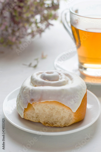 A bun covered with white glaze on a saucer, a cup of tea and a flower in a pot. Baking with tea in the morning light.