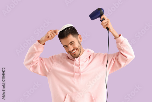 Handsome young man with hair dryer and brush on lilac background