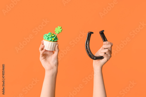 Female hands with tasty cupcake and horseshoe for St. Patrick's Day on orange background