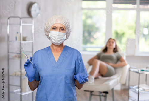 Portrait of trustworthy sneior doctor in scrub and mask holding syringe with patient lying in the back.