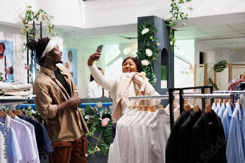 African american woman customer taking selfie on smartphone while choosing clothes in shopping mall. Fashion boutique clients trying formal wear and making photo on mobile phone