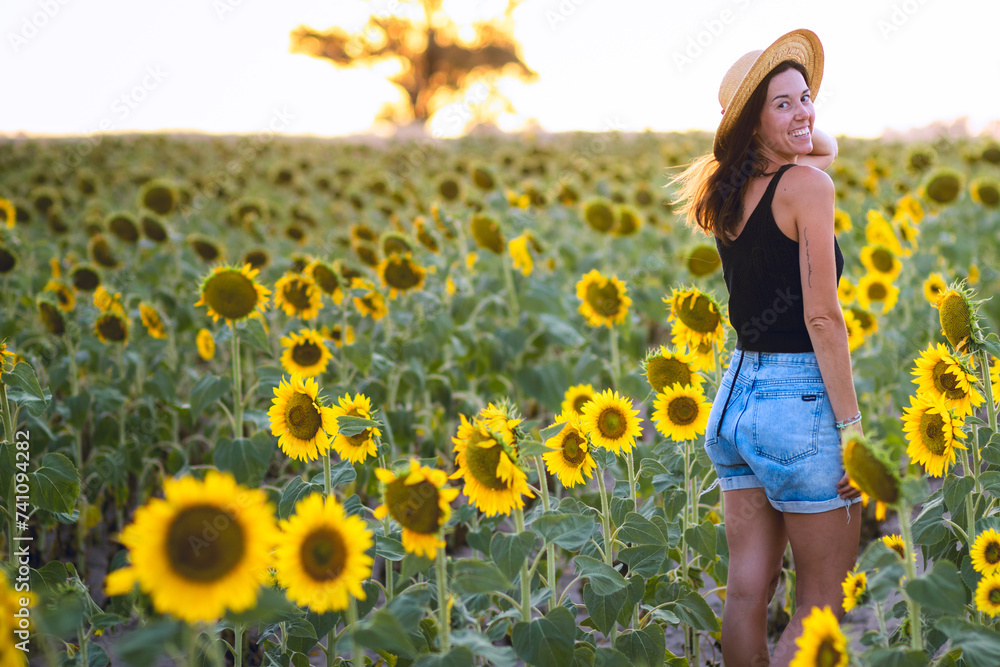 Young woman from behind looking at camera in a sunflower plantation during golden hour