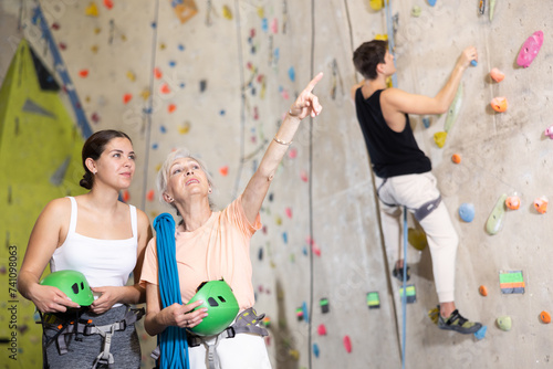 two active female marathon participants are standing in climbing hall. elderly woman points with her hand towards wall with difficult climbing track