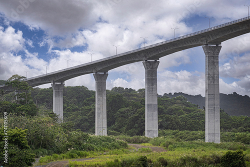 Panama Canal, Panama - July 24, 2023: Atlantic Bridge modern pillar construction just out of canal, in green forested landscape under blue cloudscape